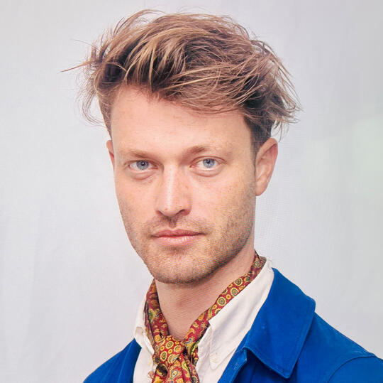 Blonde, white man with grey eyes looking into the camera, not smiling. Wearing a blue blazer, white collared shirt and a green ascot with yellow and green circles.