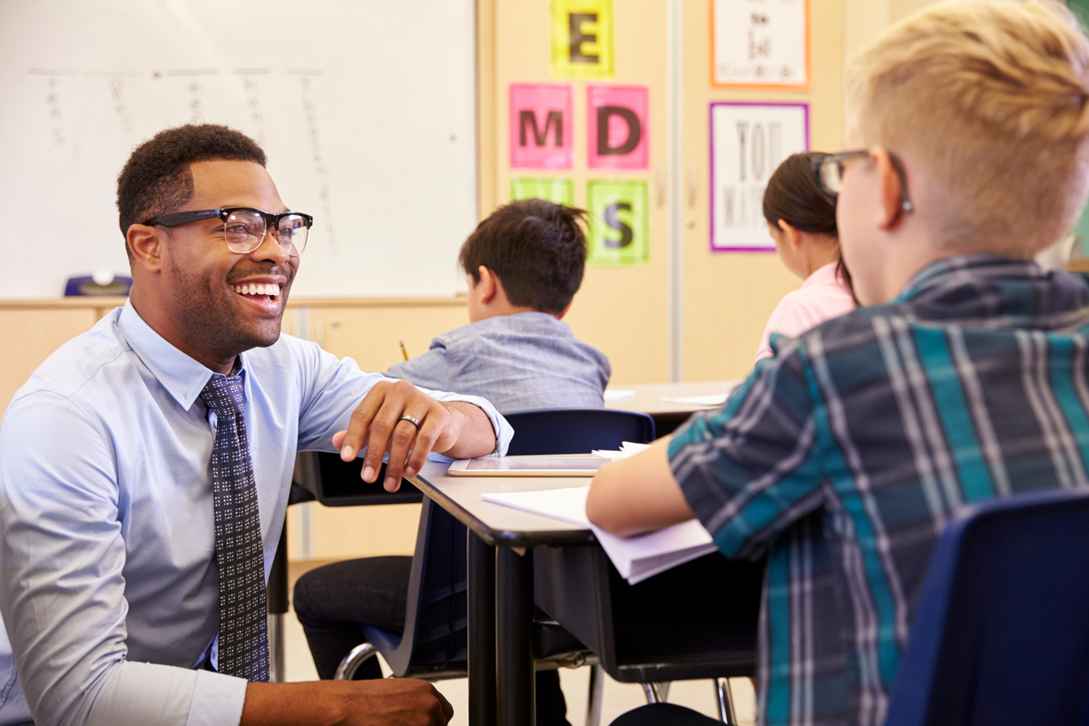 A teacher kneels beside an elementary student's desk and smiles