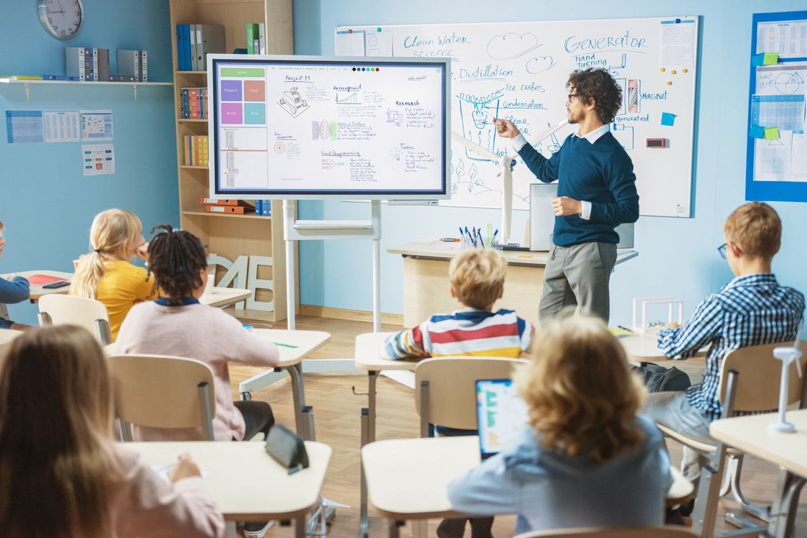 Teacher in a classroom pointing at a presentation on a screen, teaching a class of diverse students