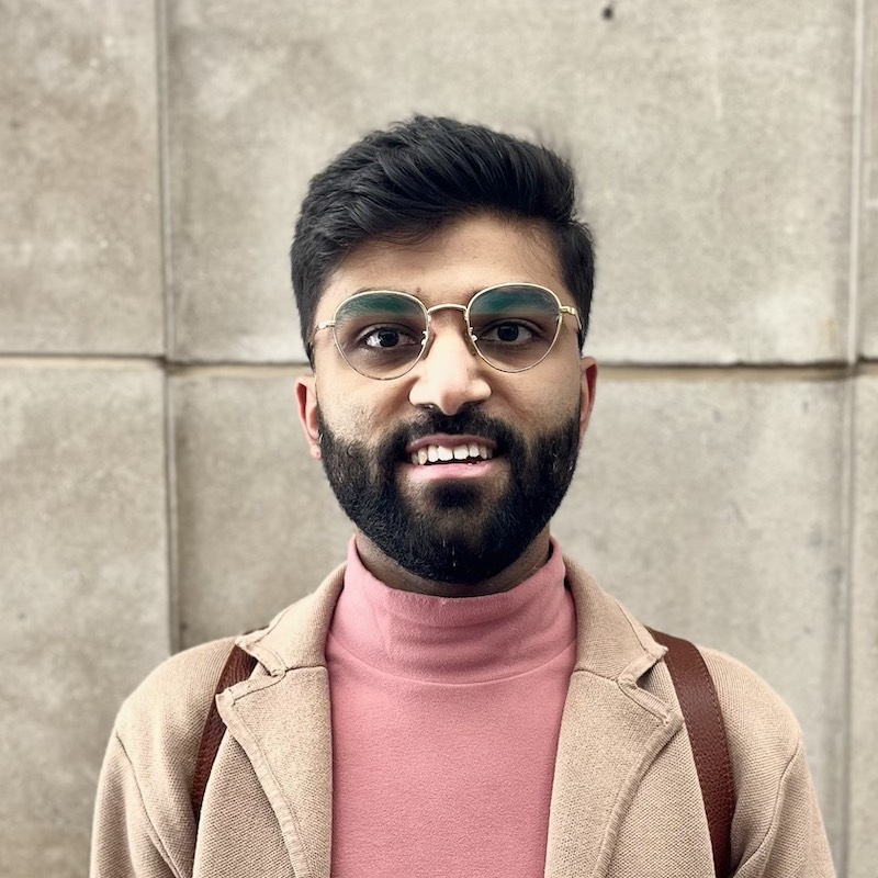 Raaghav Pandya smiling in front of a stone wall. He has a full beard and gold rimmed glasses.