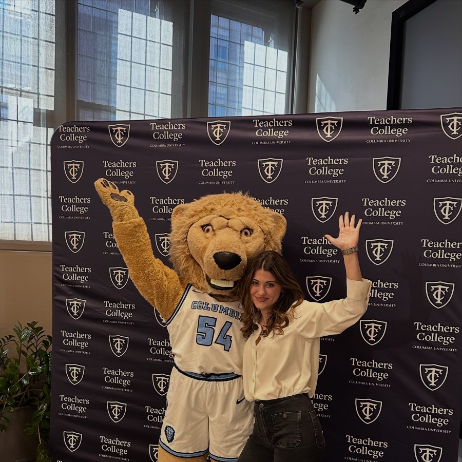 A student standing in front of a Teachers College branded step and repeat with the Columbia University mascot