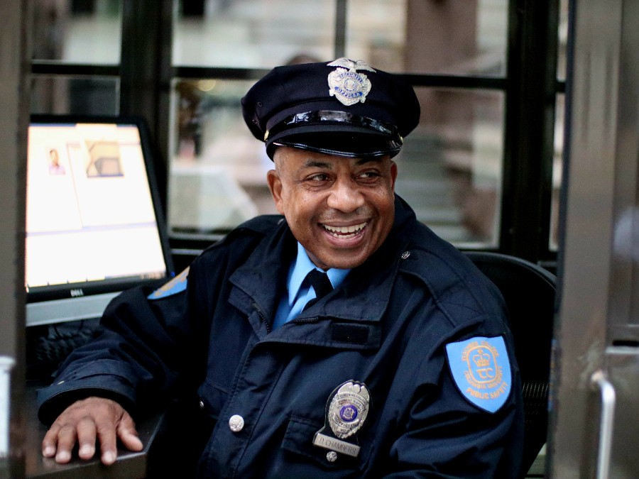 Dr. Dennis Chambers, a smiling Black man, sits inside a security booth wearing a dark blue public safety officer uniform complete with a peaked cap, silver chest badge, nameplate, and shoulder patch. He rests one hand on the desk next to a computer monitor and looks off-camera with a warm, cheerful expression.