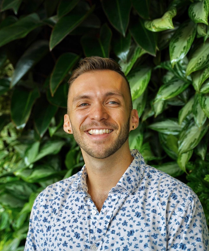 A photograph taken against a background of green foliage of a smiling Joey Haavik (he/him)