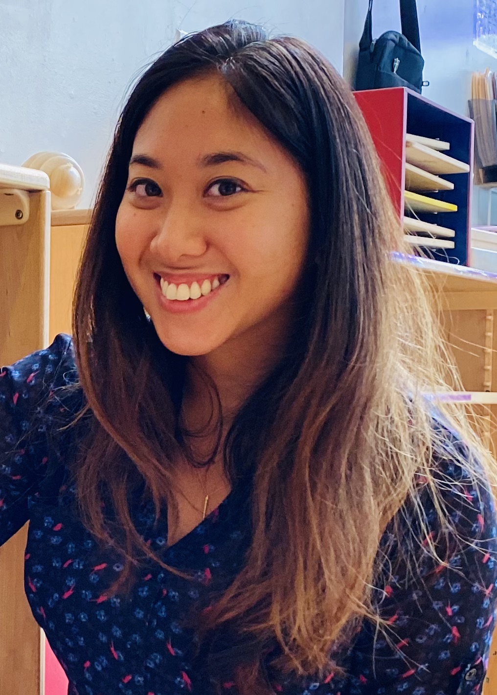 A woman with long brown hair smiling and looking at the camera. She is wearing a dark blue blouse with a light pink floral pattern. The background is a classroom.