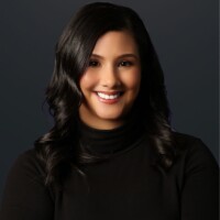A headshot of Michele Lopez, a smiling woman with dark, shoulder-length wavy hair, looking at the camera. She is wearing a black turtleneck against a solid dark gray background.