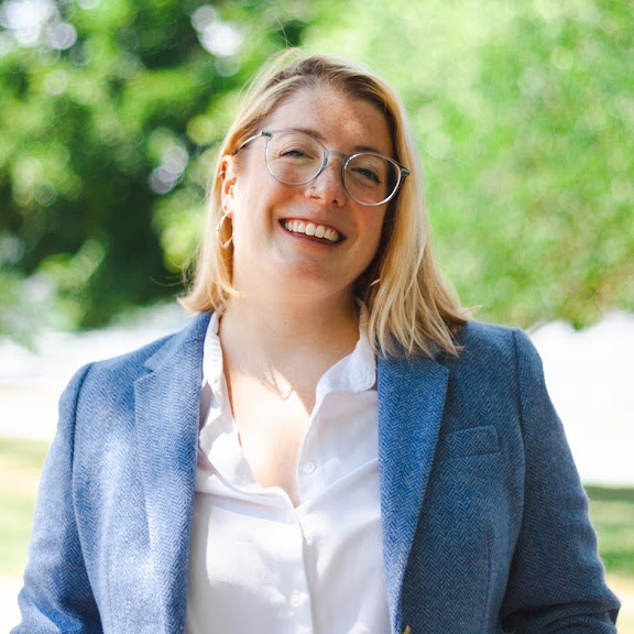 A woman with shoulder-length blonde hair, wearing a white shirt and a blue blazer. She is smiling and looking to her left. She is wearing round glasses and has a nose ring. The background is blurred with green trees.