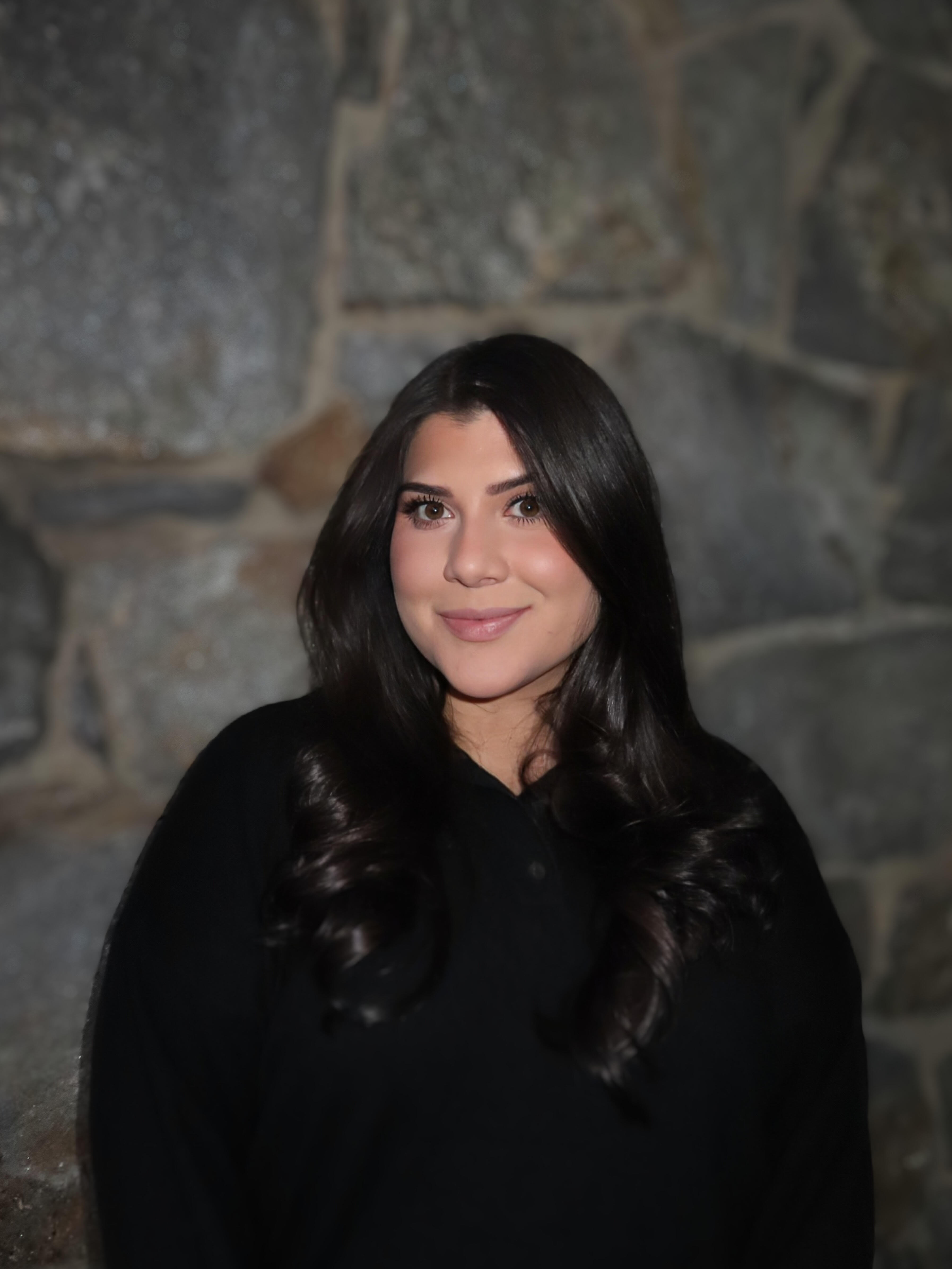 A professional headshot of a smiling young woman with long, dark brown hair. She is wearing a black collared shirt and is standing in front of a stone wall.
