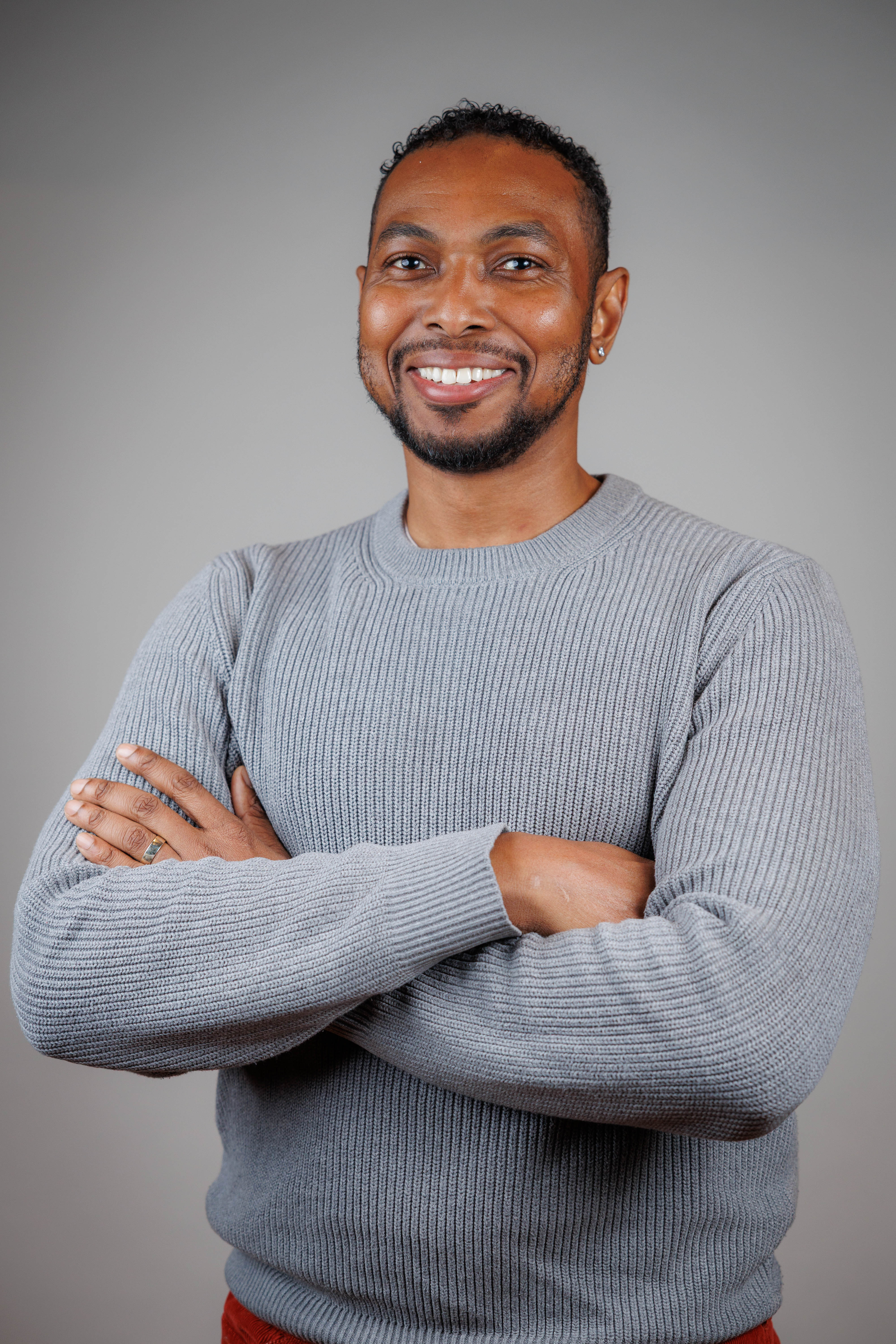 A headshot of a smiling Black man with a goatee and short dreadlocks. He is wearing a gray ribbed sweater and has his arms crossed.