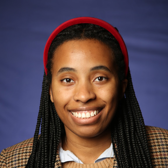 A professional headshot of a smiling young Black woman with long box braids and a red headband. She is wearing a brown and tan houndstooth jacket and a light blue collared shirt.