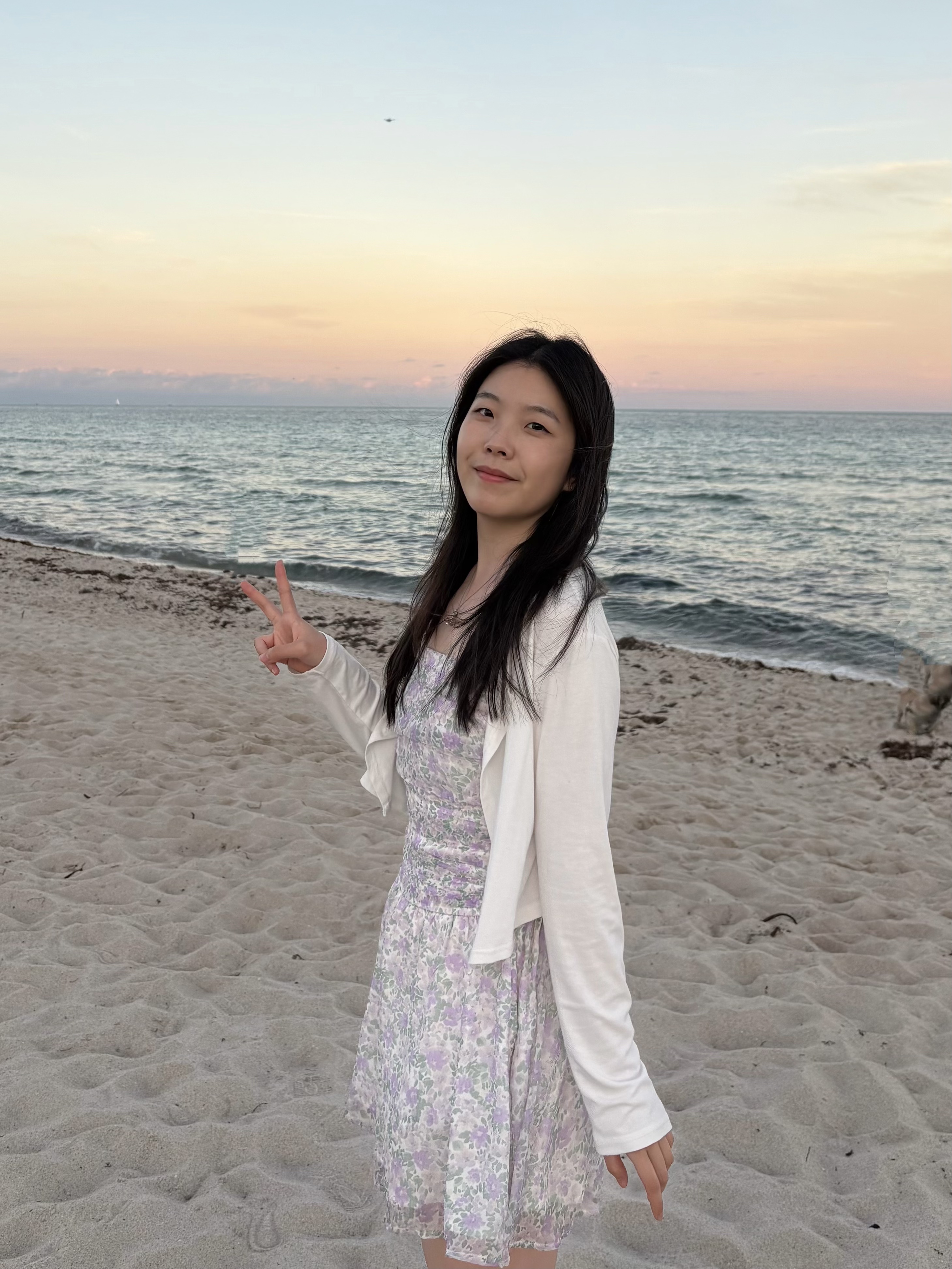 A young woman with long dark hair stands on a sandy beach at sunset, making a peace sign with her hand. She is wearing a floral print sundress in shades of purple and white, paired with a white long-sleeved cardigan. The background features a calm ocean under a soft gradient sky of orange, pink, and light blue.