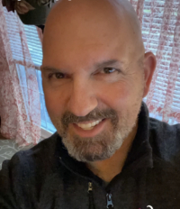 A close up photo of a smiling man with a gray goatee wearing a black collared shirt.
