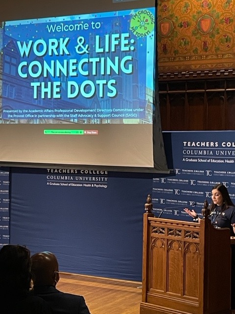 Photo of a presentation projected on screen, and an individual speaking at a podium in a gothic wooden auditorium.