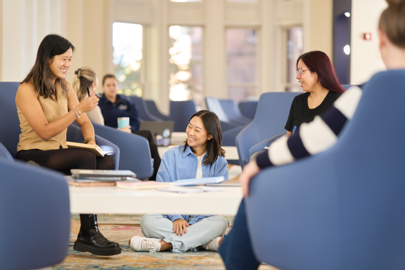 A diverse group of college students relax and chat in a bright, modern lounge. Three women are in the foreground, with one seated on the floor and two in blue armchairs, engaged in conversation and smiling. Others are visible in the background.