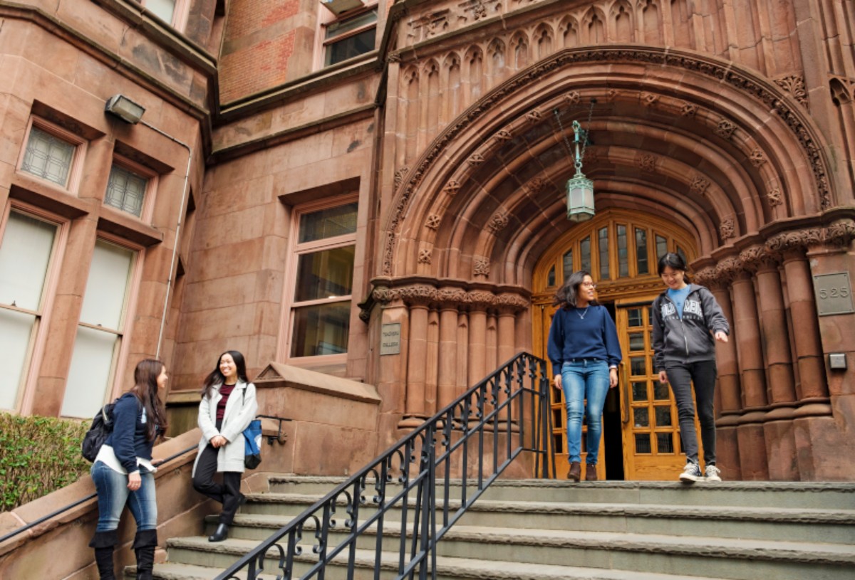 Four diverse students are walking up and down the stone steps of a grand, historic brownstone building with an arched doorway. Two are on the lower steps and two are near the top, by the entrance.