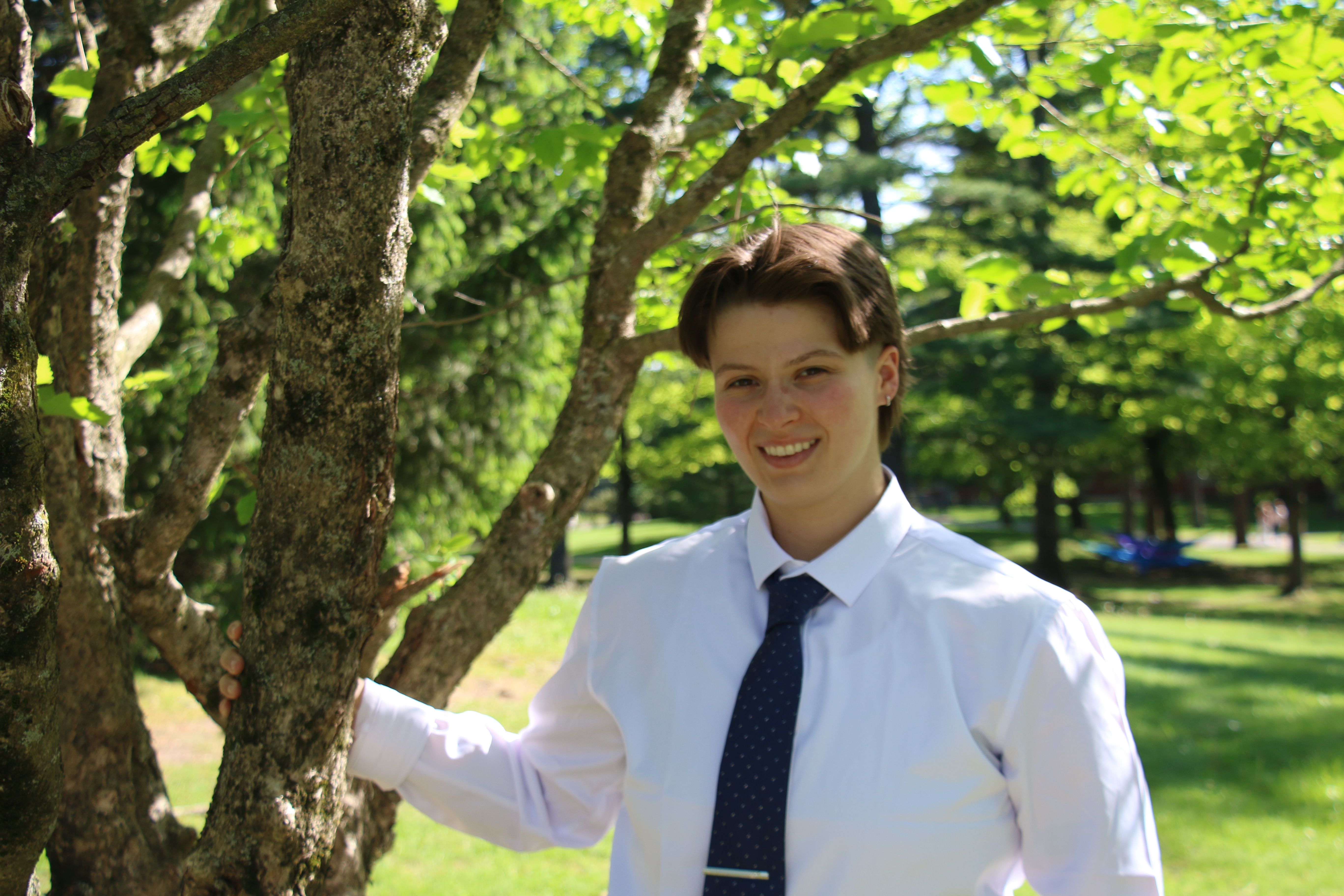 Photo of student wearing a white shirt and dark tie standing in front of a tree