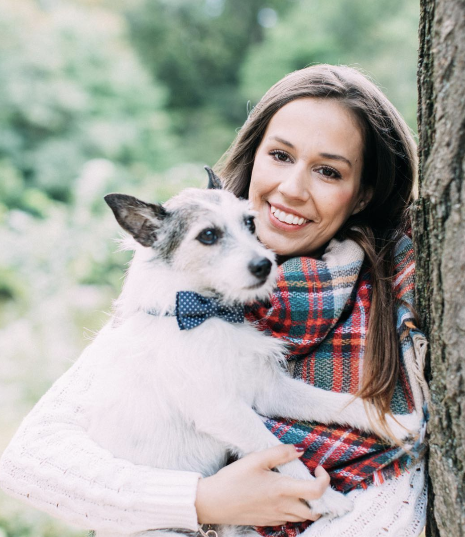 Whitney leaning against a tree in park holding small white dog with bowtie