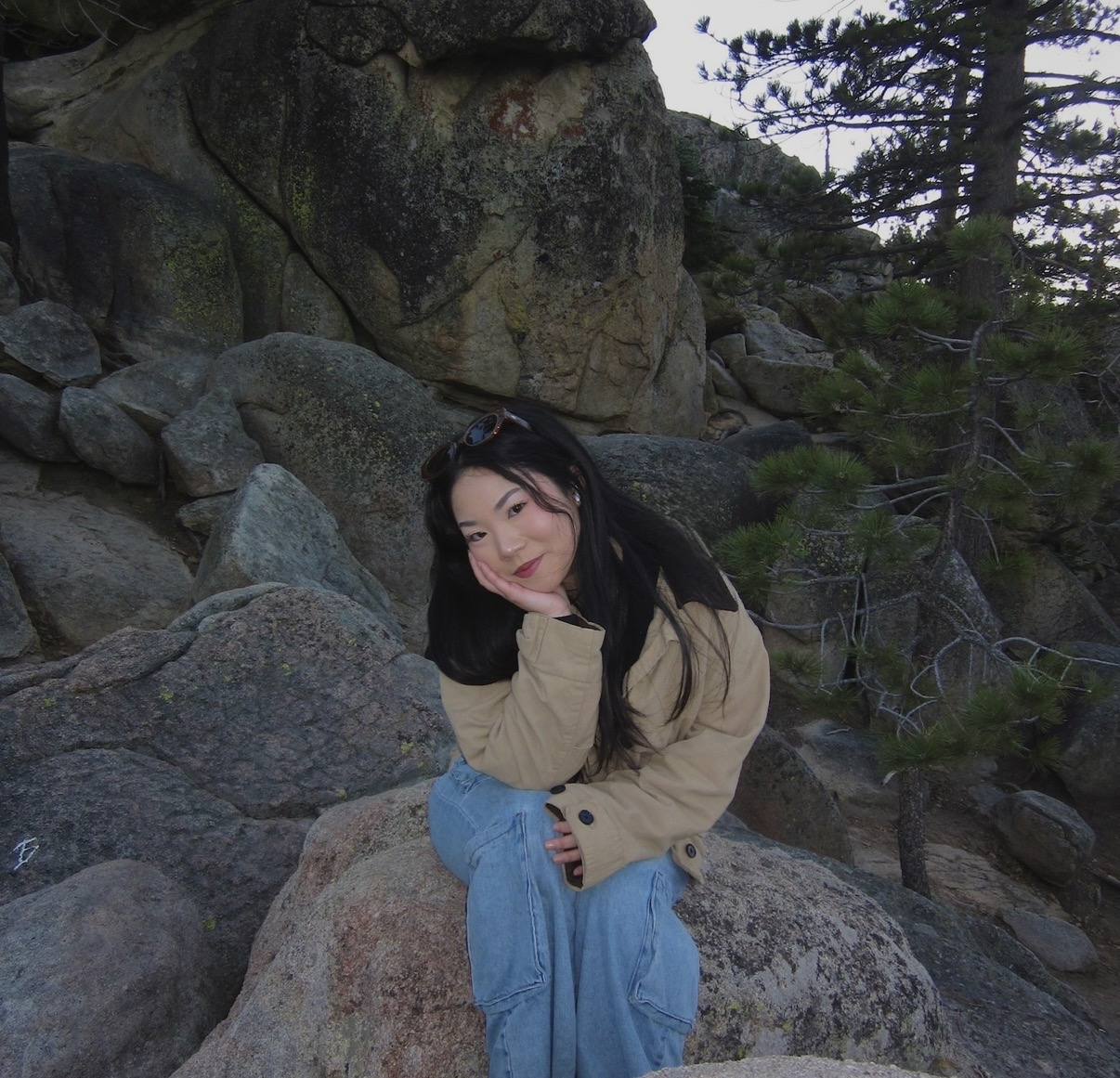 A young woman with long dark hair poses outdoors while sitting on a large boulder. She rests her chin on her hand, leaning forward slightly with a slight smile. She wears sunglasses pushed up on her head, a beige jacket, and light blue jeans. Surrounding her are large grey rock formations and evergreen pine trees.