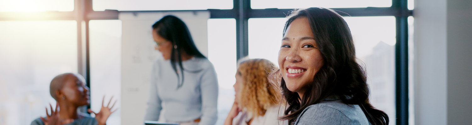 A smiling professional woman turns toward the camera during a meeting, with colleagues engaged in discussion and a presenter at a whiteboard visible in the bright, sunlit background.