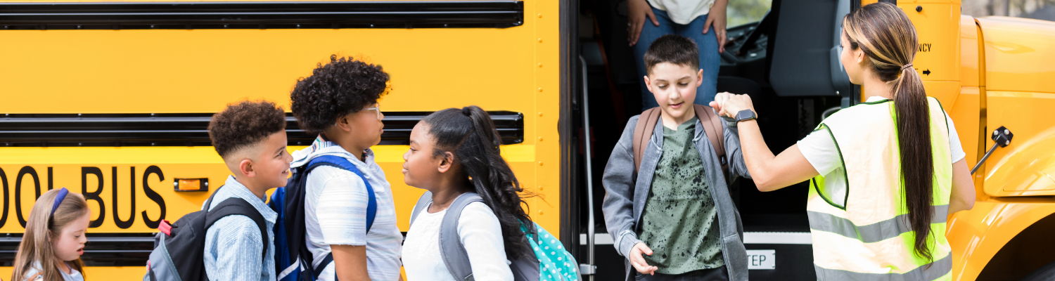 A group of elementary school children with backpacks lining up to board a yellow school bus, greeted by a staff member in a high-visibility safety vest holding the door open.