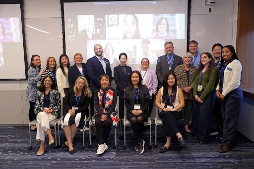 Group shot of the TC alumni council with some people standing in the background and other seated in the foreground, with a projection with faces on a screen behind them.