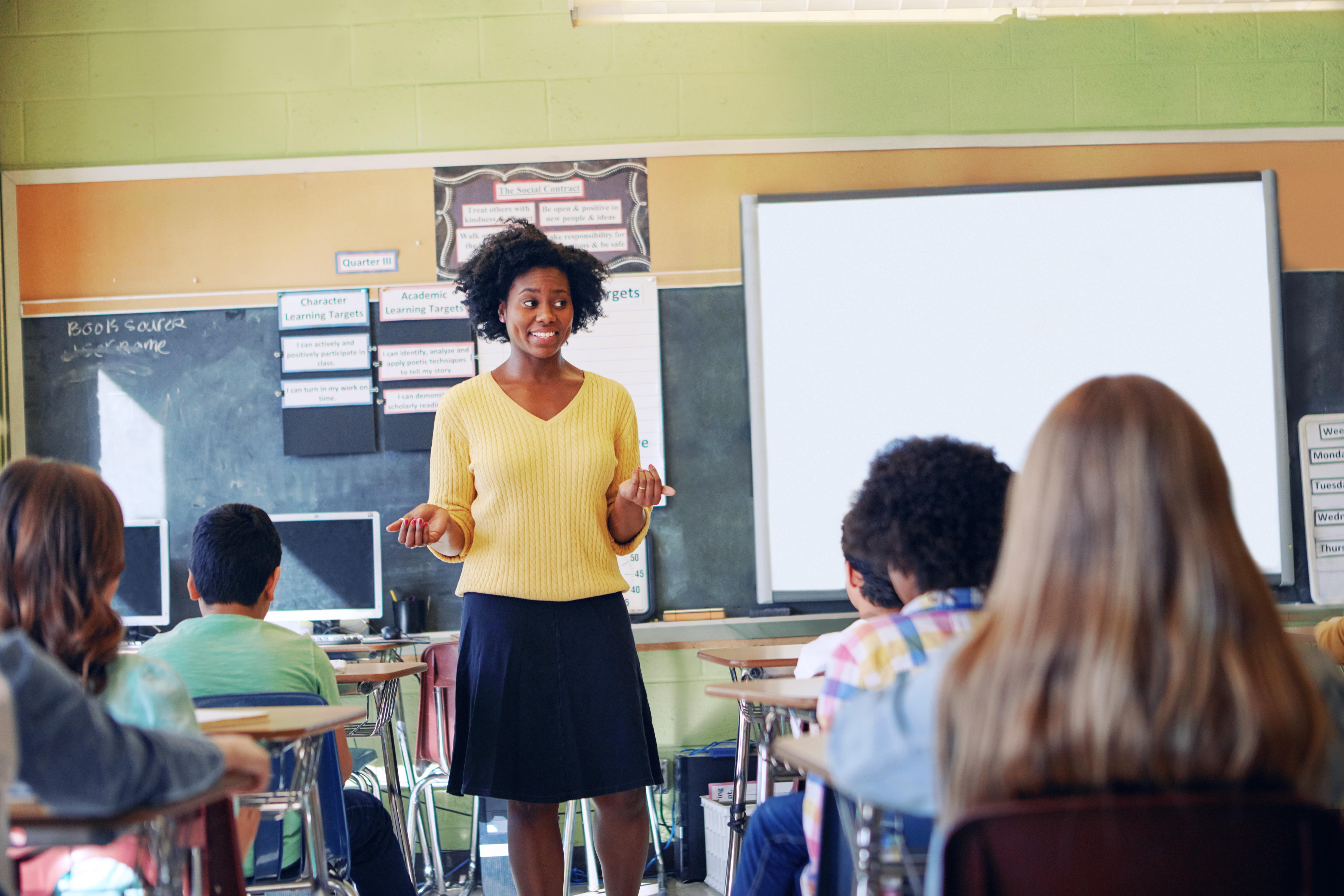 A teacher in a yellow sweater standing at the front of an elementary classroom.