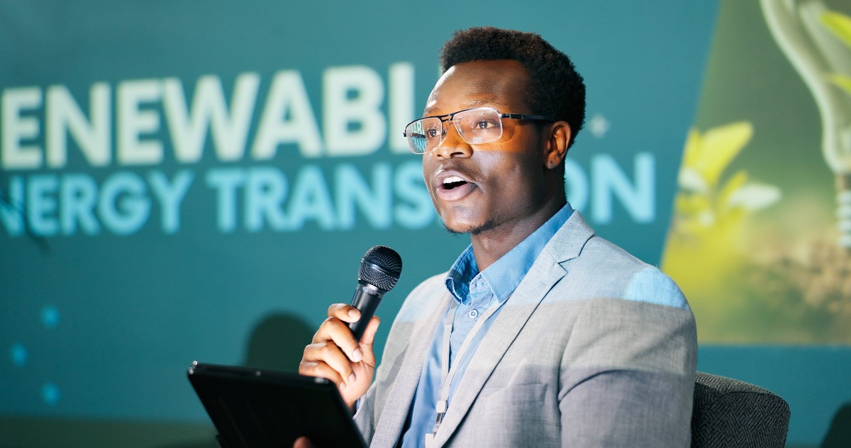 A male speaker holding a microphone and a tablet standing in front of a screen with the words renewable energy transition.