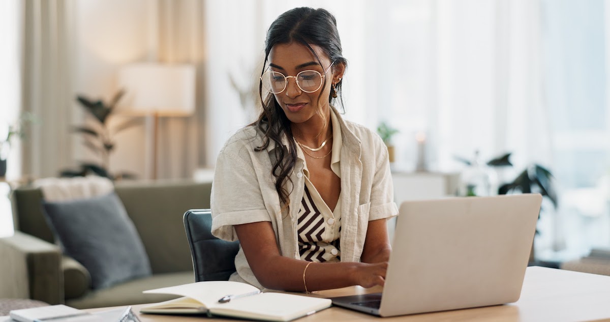 A woman with glasses sitting at a table looking at an open notebook while typing on a laptop