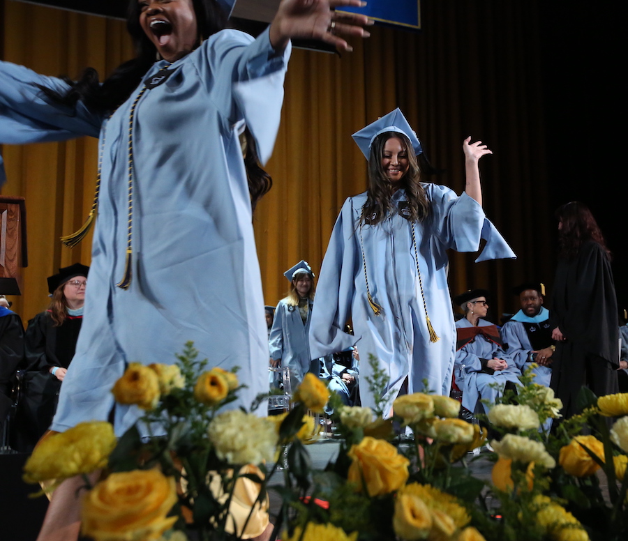 Two students wearing graduation regalia walk across the stage