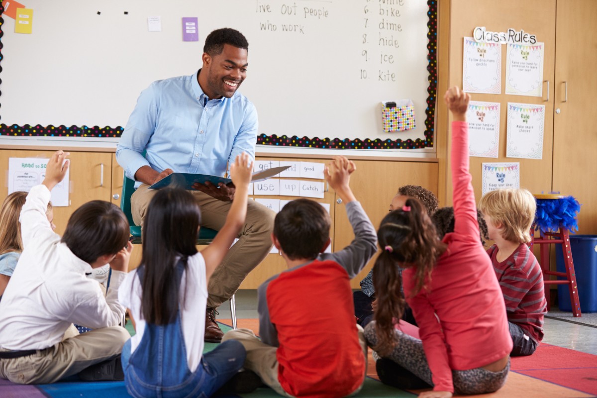 One teacher sitting at the front of a classroom with a book open in their lap. Children sit in a semi-circle in front of them.