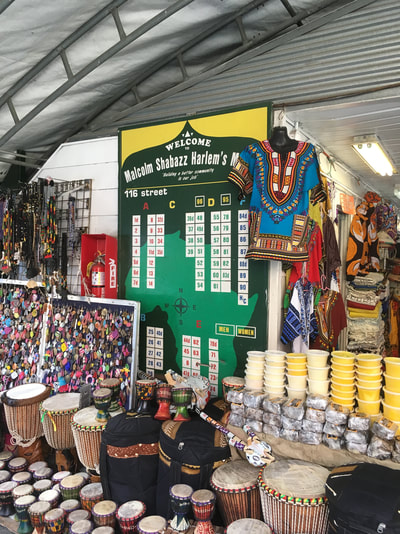 Welcome to Malcolm Shabazz Harlem's Market board with Africa in the background with clothing, drums, and various goods surrounding the board outside a shop.