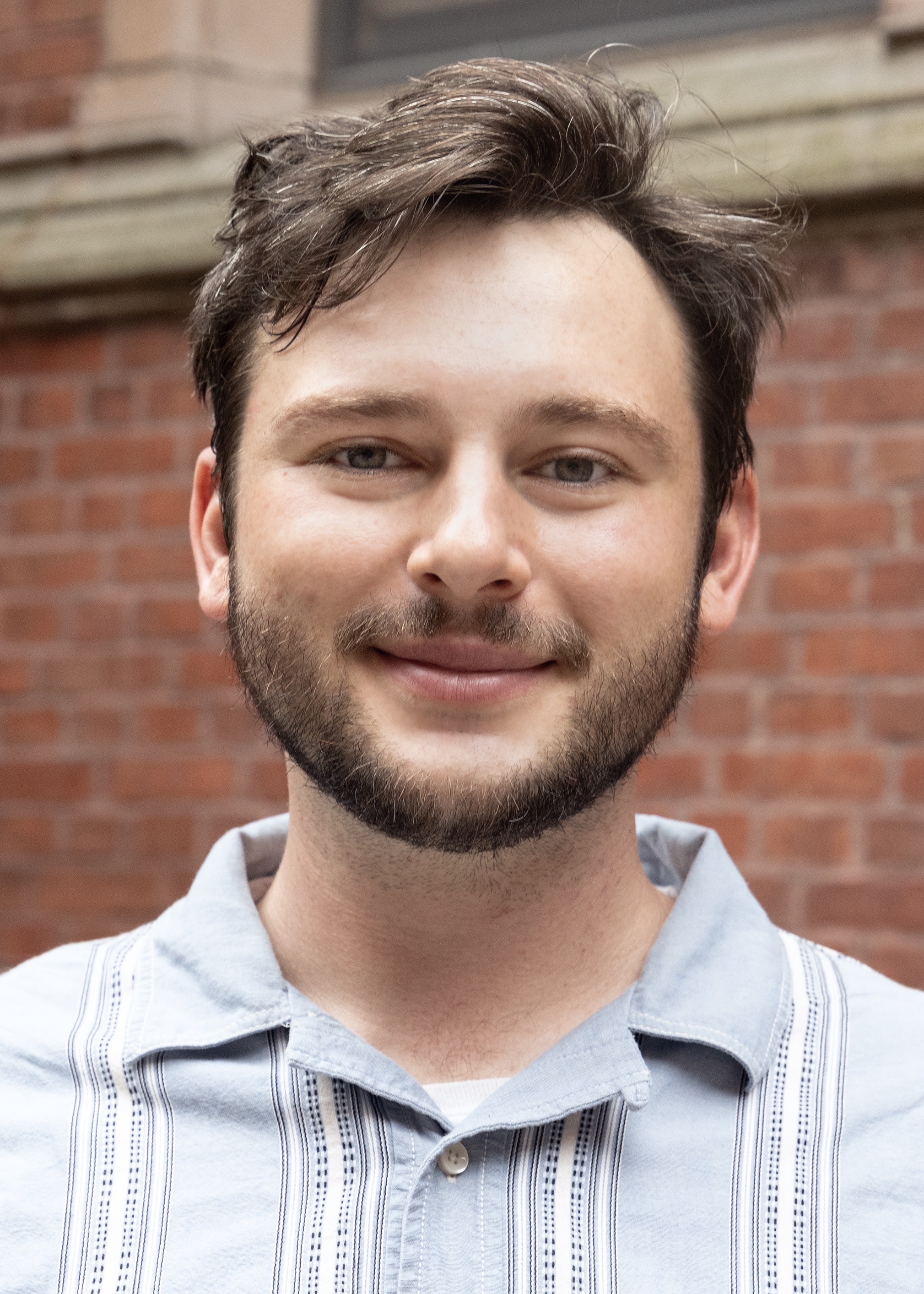 Chris smiles at the camera. He has dark blond hair and is wearing a light blue button up shirt.