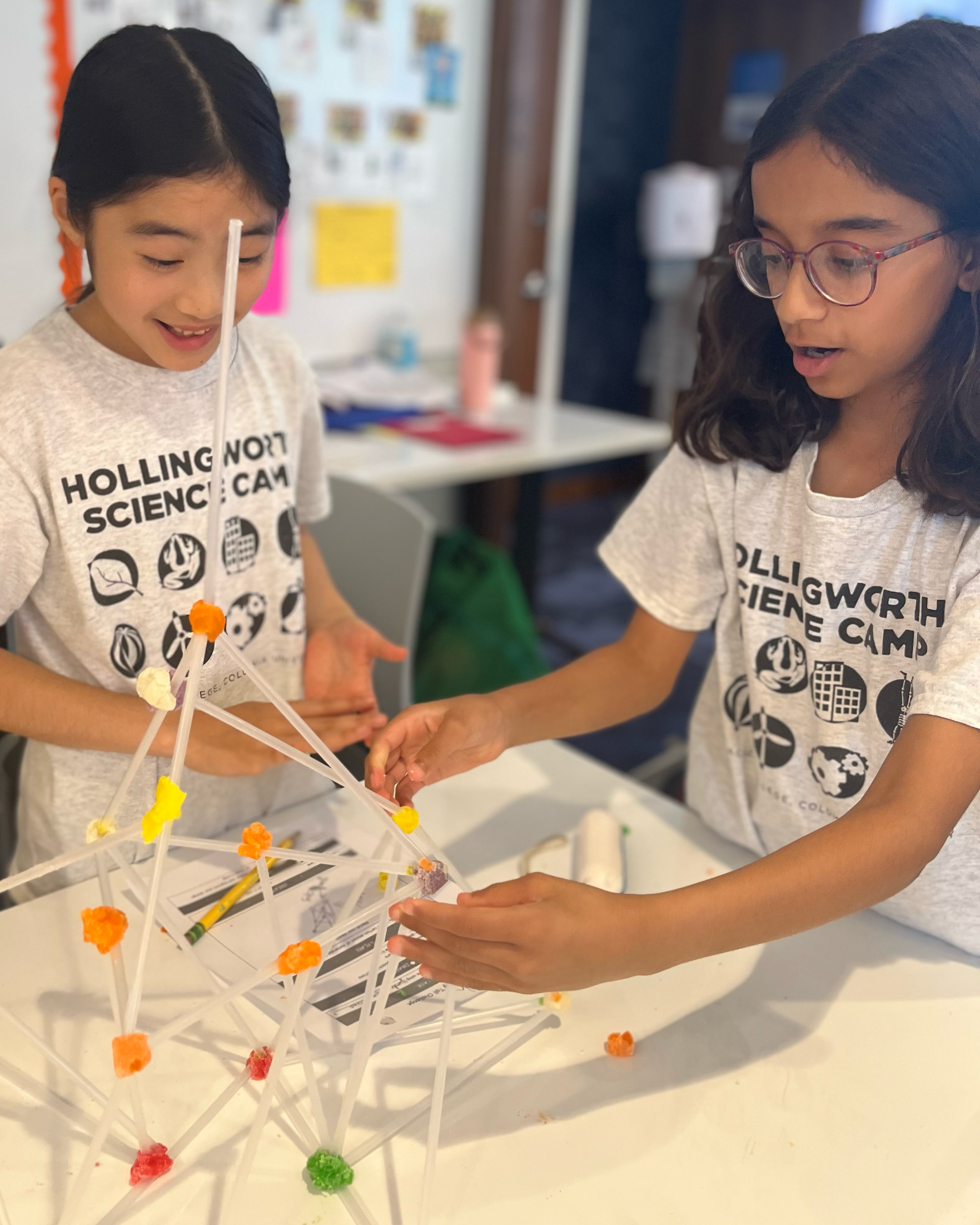 Two young girls are working together to build with toothpicks and gumdrops. They are smiling as they work urgently to keep the fragile structure standing.