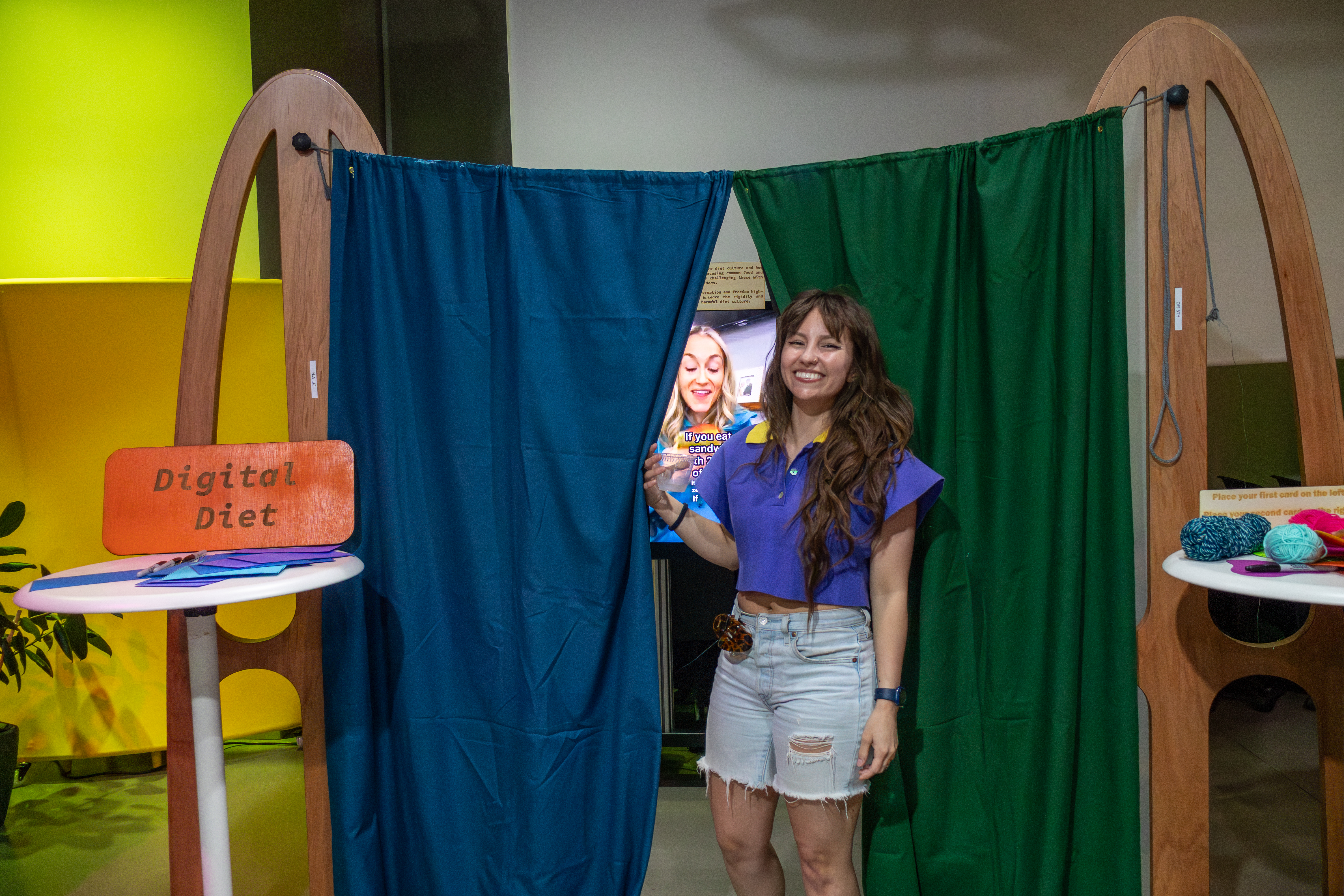 A woman smiling and standing in front of a makeshift curtain in between two tables. One table has a sign reading Digital Diet. The split curtain reveals a screen in the background.