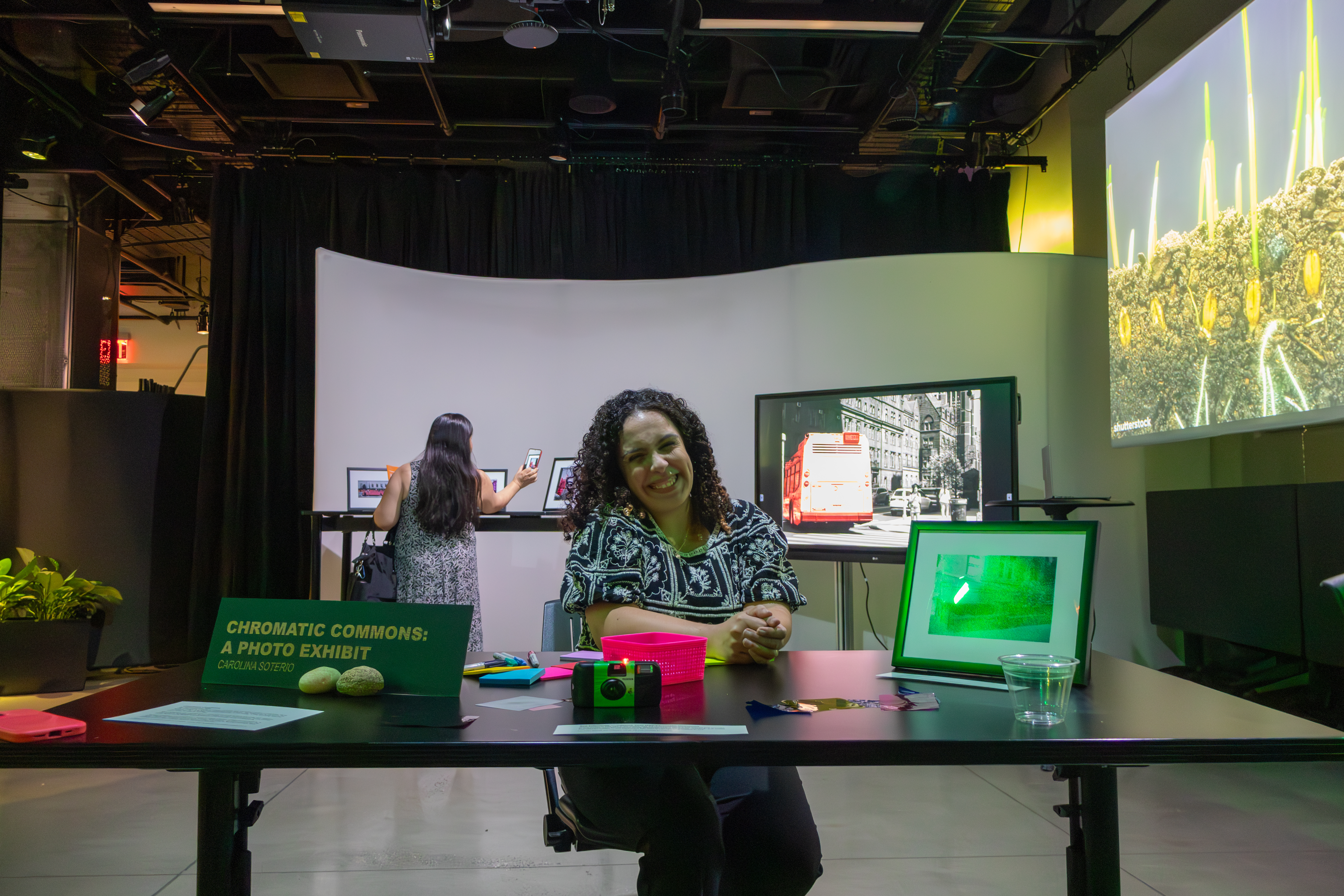 A woman sitting at a table in an exhibit room with a sign Chromatic Commons: A Photo Exhibit. The table has a disposable camera and colorful post it notes on it.