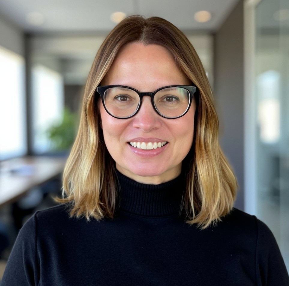 Headshot of a smiling woman with shoulder-length light brown hair wearing black glasses and a black turtleneck, standing in a bright office hallway