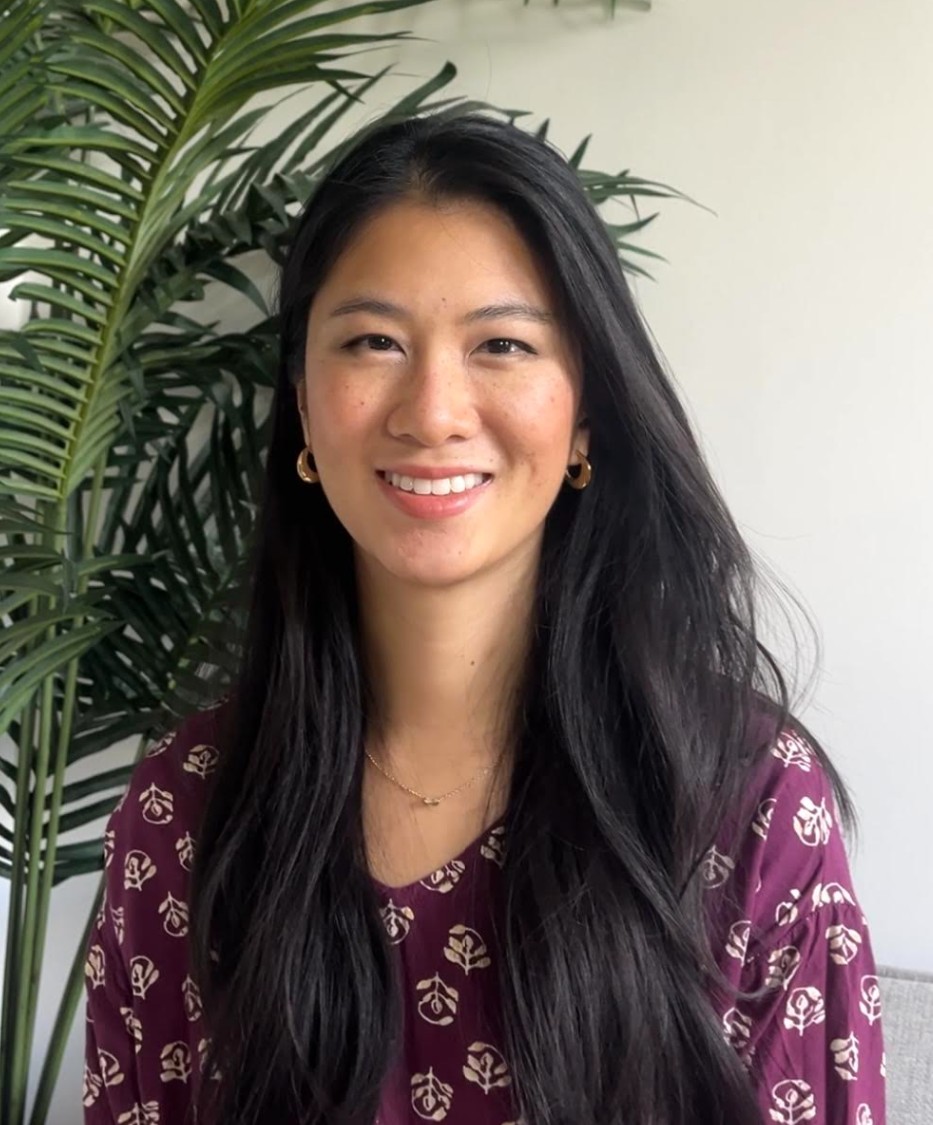 A professional headshot of a woman smiling while seated indoors. She is wearing a patterned maroon top and small gold hoop earrings, with a green plant visible in the background