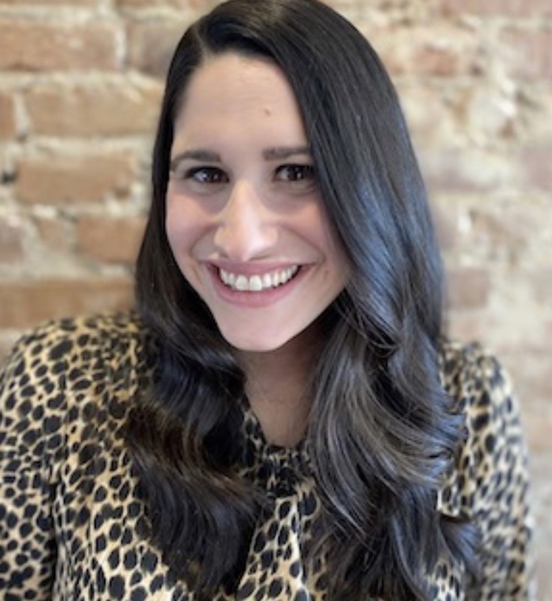 A woman with long dark hair smiles at the camera, wearing a patterned leopard top, with a brick wall in the background.