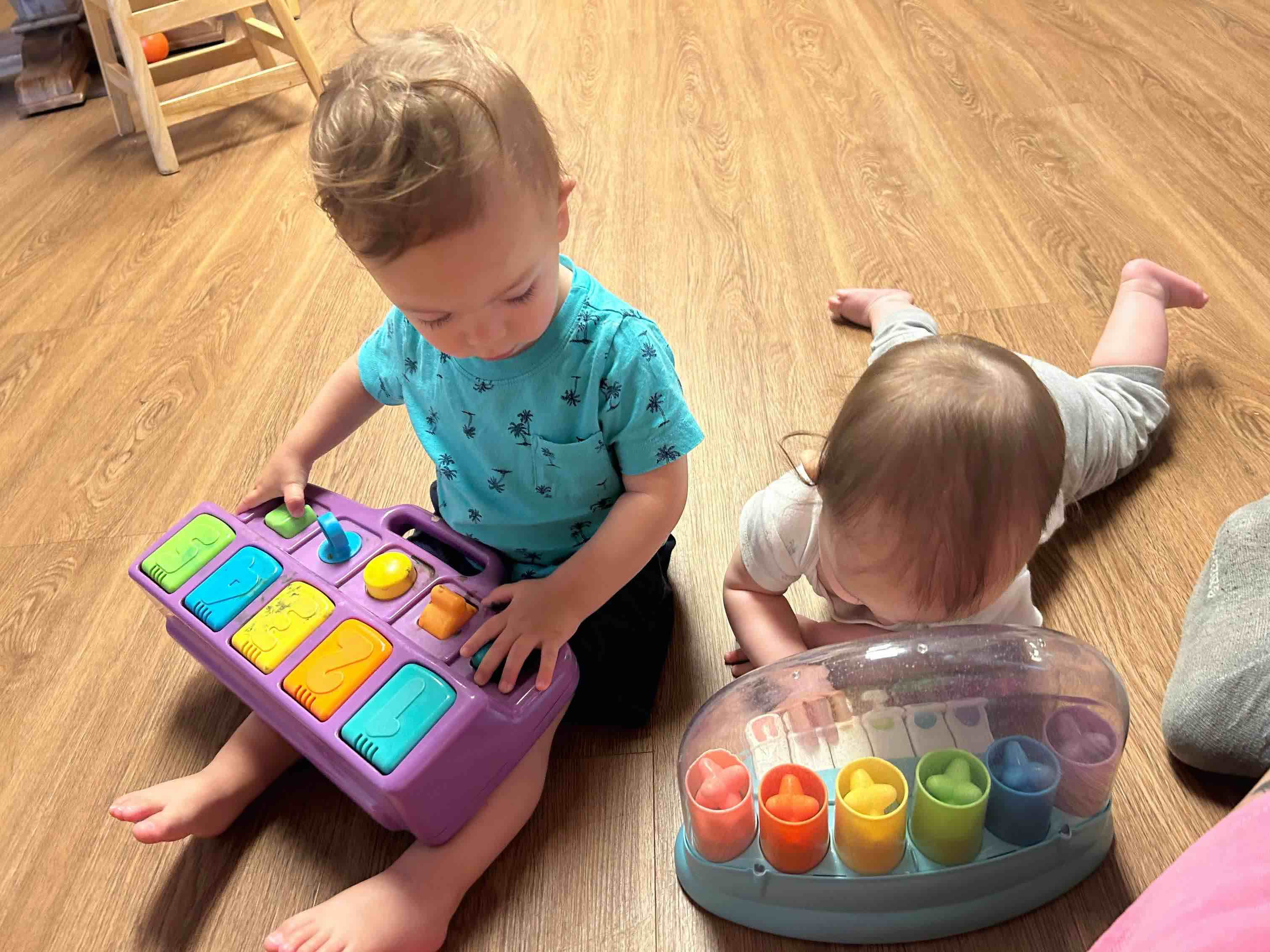 An infant on her tummy plays with a toy next to a toddler playing with a toy on his lap