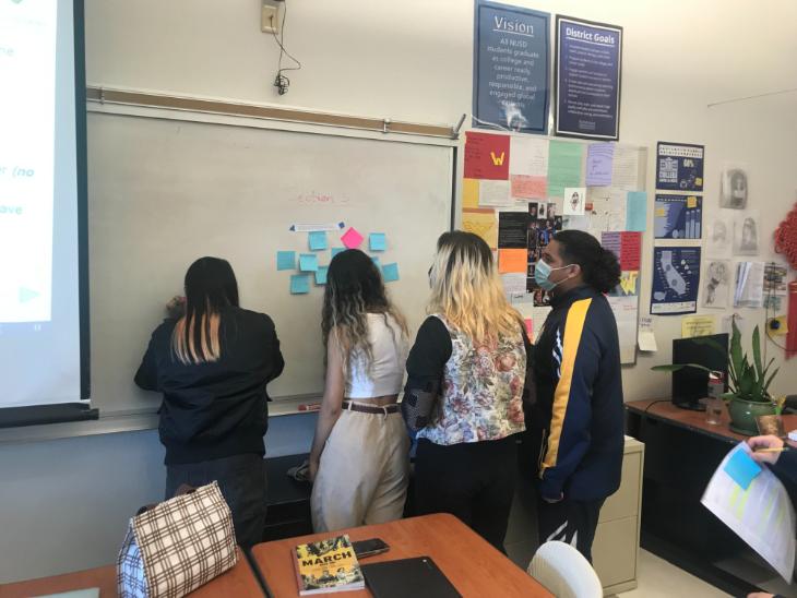 Four high school students cluster around a white board in a classroom, writing on post-it notes.