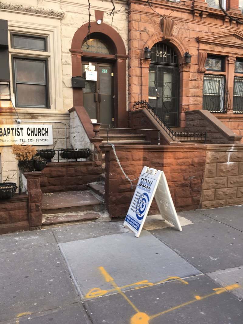 Brownstone walk-up with white foldable sign in front and a white Baptist Church sign on the building