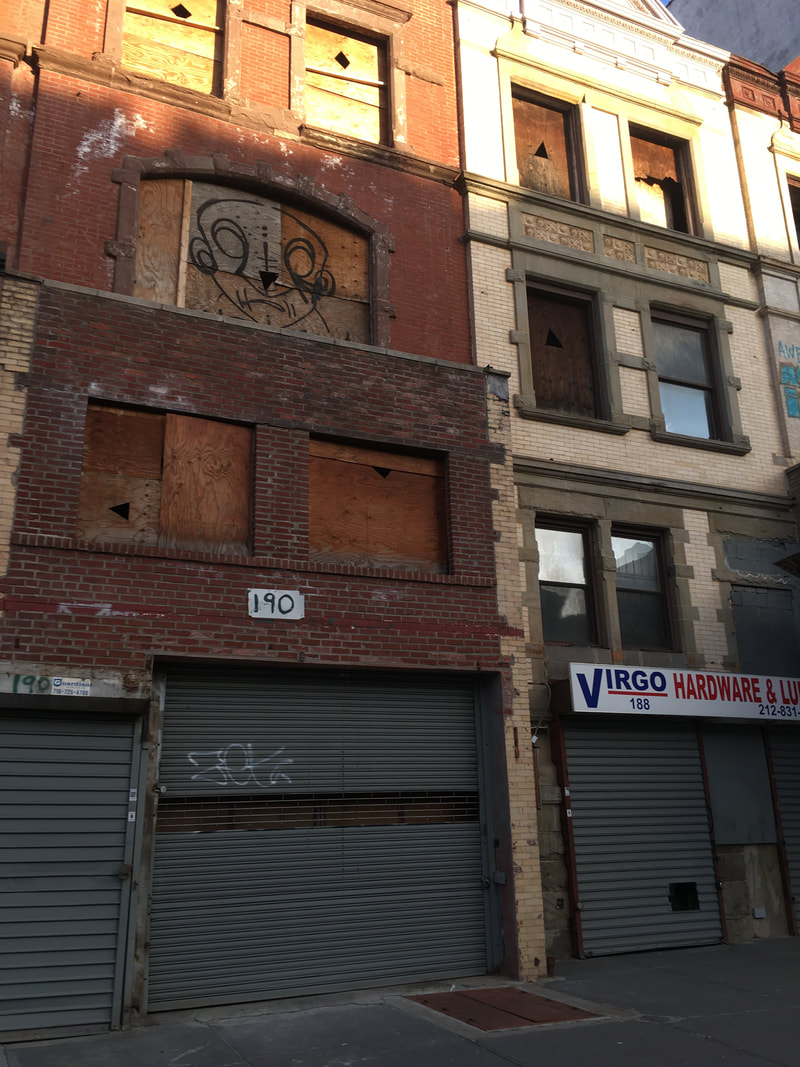 Boarded windows of a red brick home next to a tan walk-up with glass windows and closed storefronts below