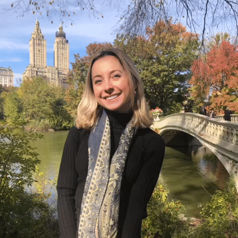 Tay smiles at the camera. She is in Central Park, standing in front of a bridge with trees and iconic Upper West Side buildings in the background