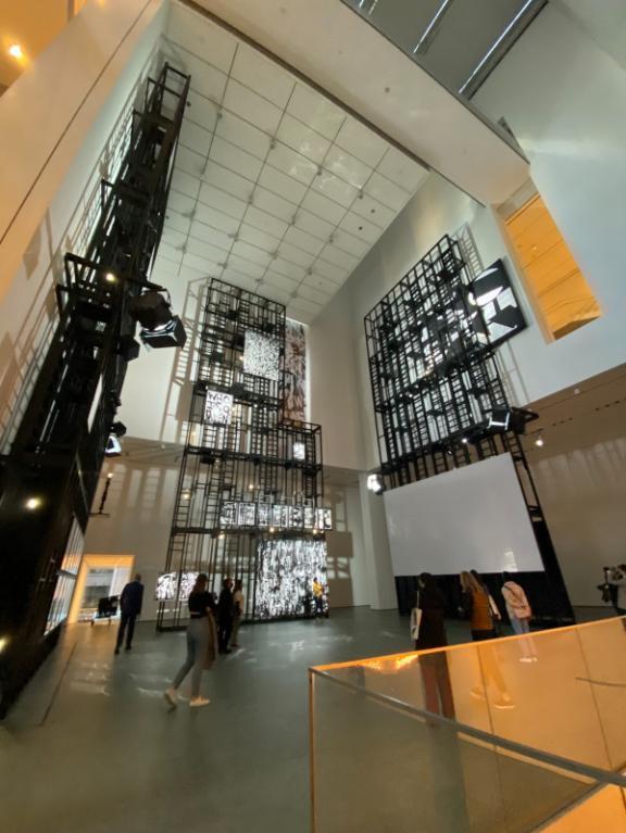 a floor-level view of Pendleton's exhibition in the 2nd floor lobby at MoMA. Tall, scaffolding-like structures extend five floor to the ceiling with projected video and large, graffiti-style paintings