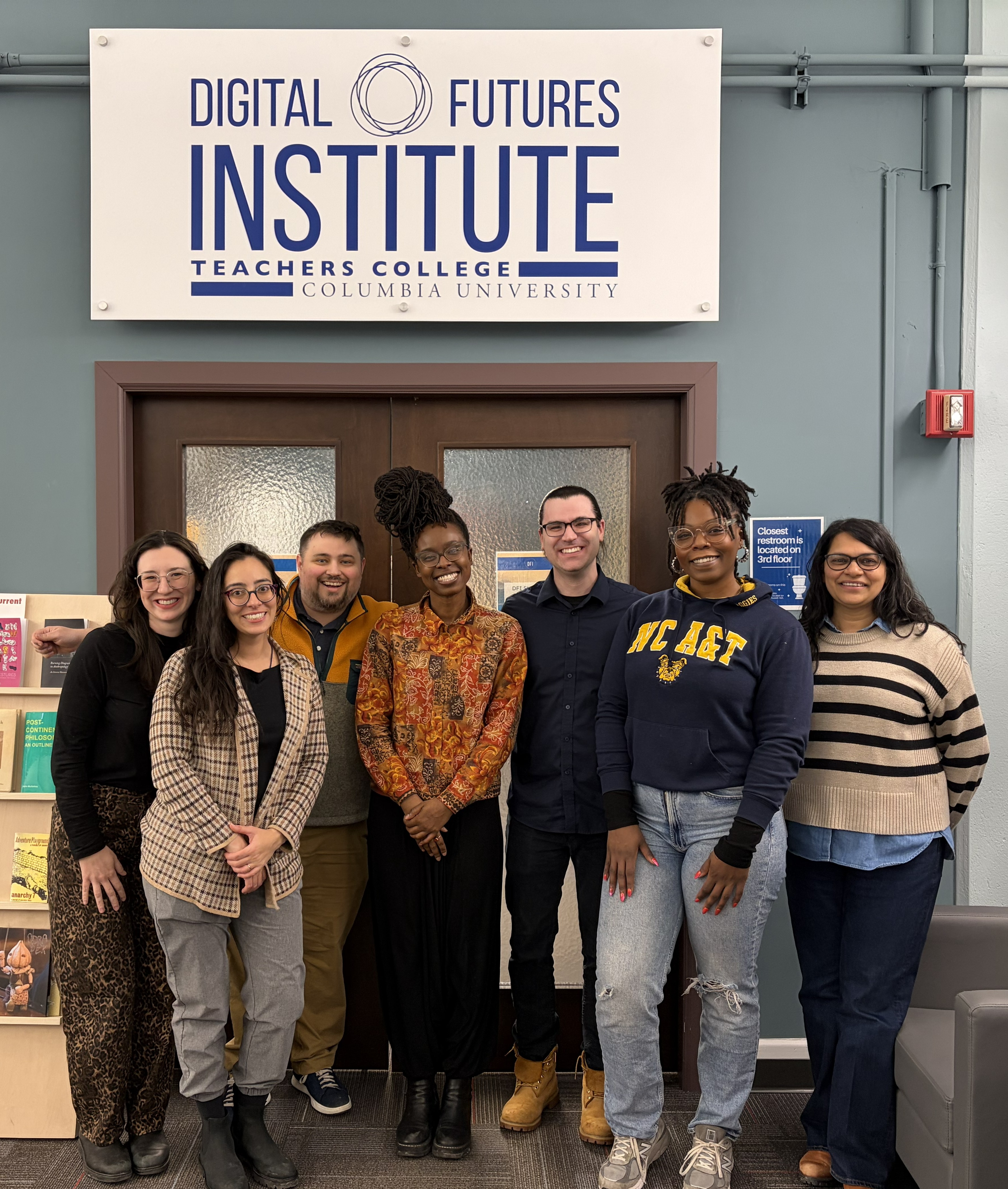 This photos shows 7 people standing together and smiling, in front of the office doors of the Digital Futures Institute (DFI). Read caption for more details.