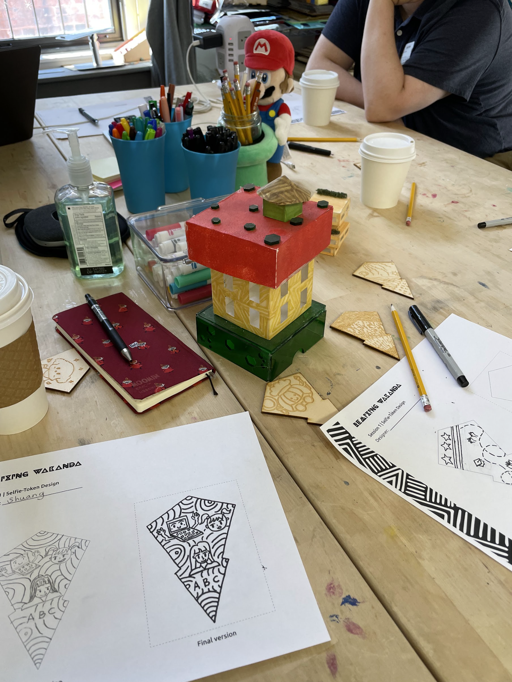 This photo shows the setting of an ERIN Fellows Constructionism and Play workshop led by Dr. Nathan Holbert. This shows a wooden table full of pens, markers, pencils, paper constructions, Remixing Wakanda Selfie Token Design pages, and coffee cups.