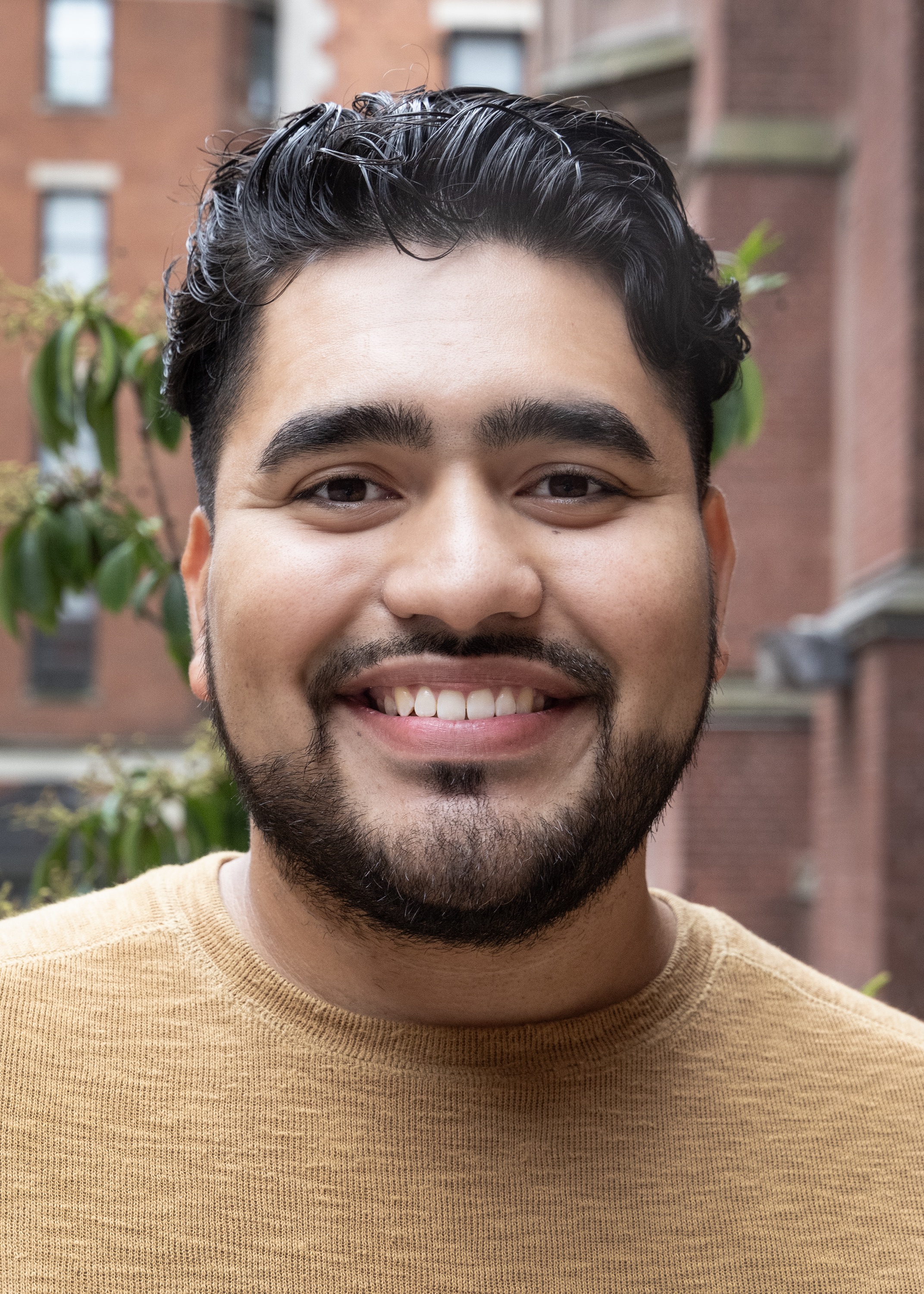 Alex is smiling at the camera. He has short, dark wavy hair and is wearing a tan sweater.