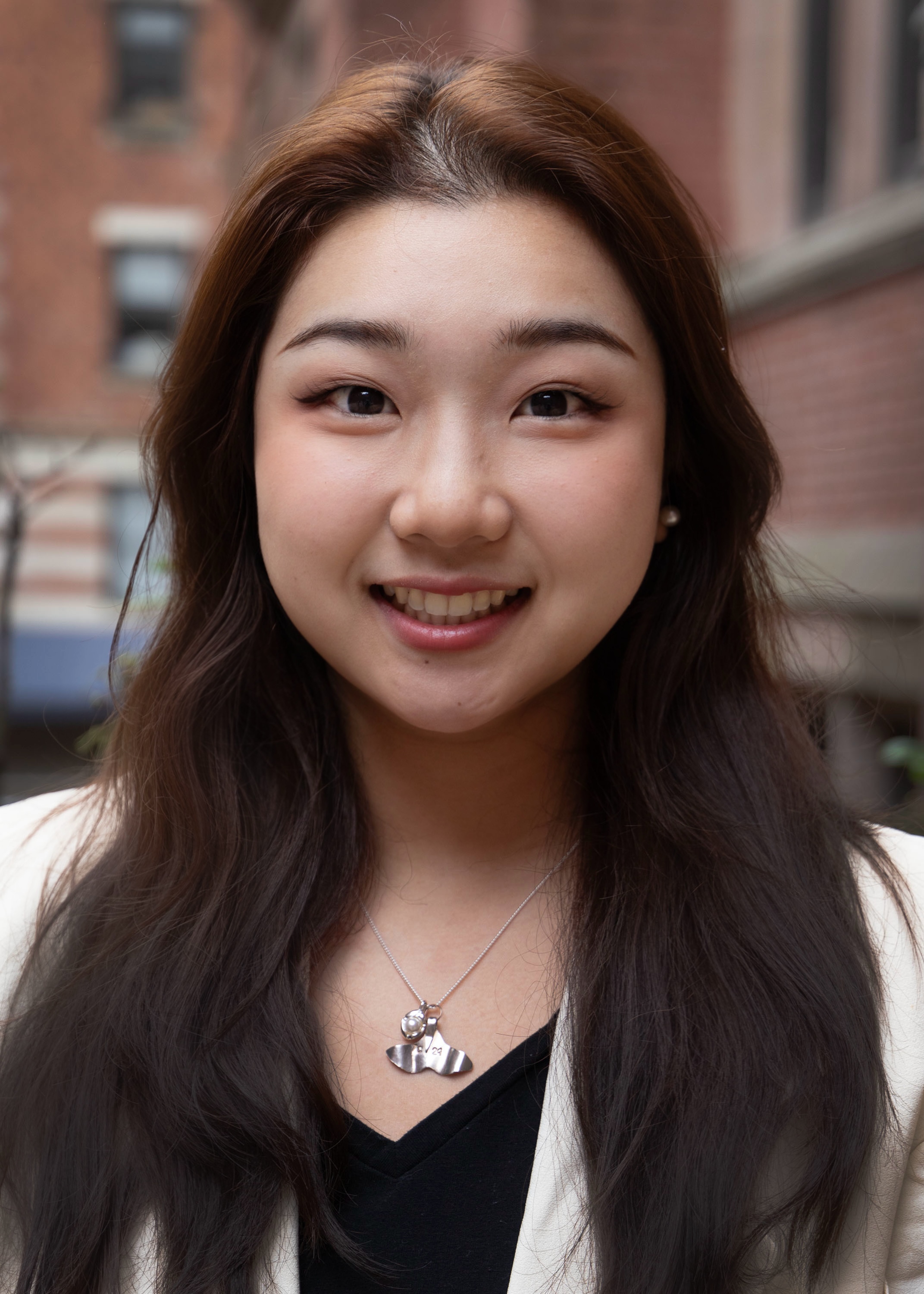 Tianxin is posing for a picture. She has long, straight, dark brown hair and a smile on her face. She is wearing a black top and off-white blazer.