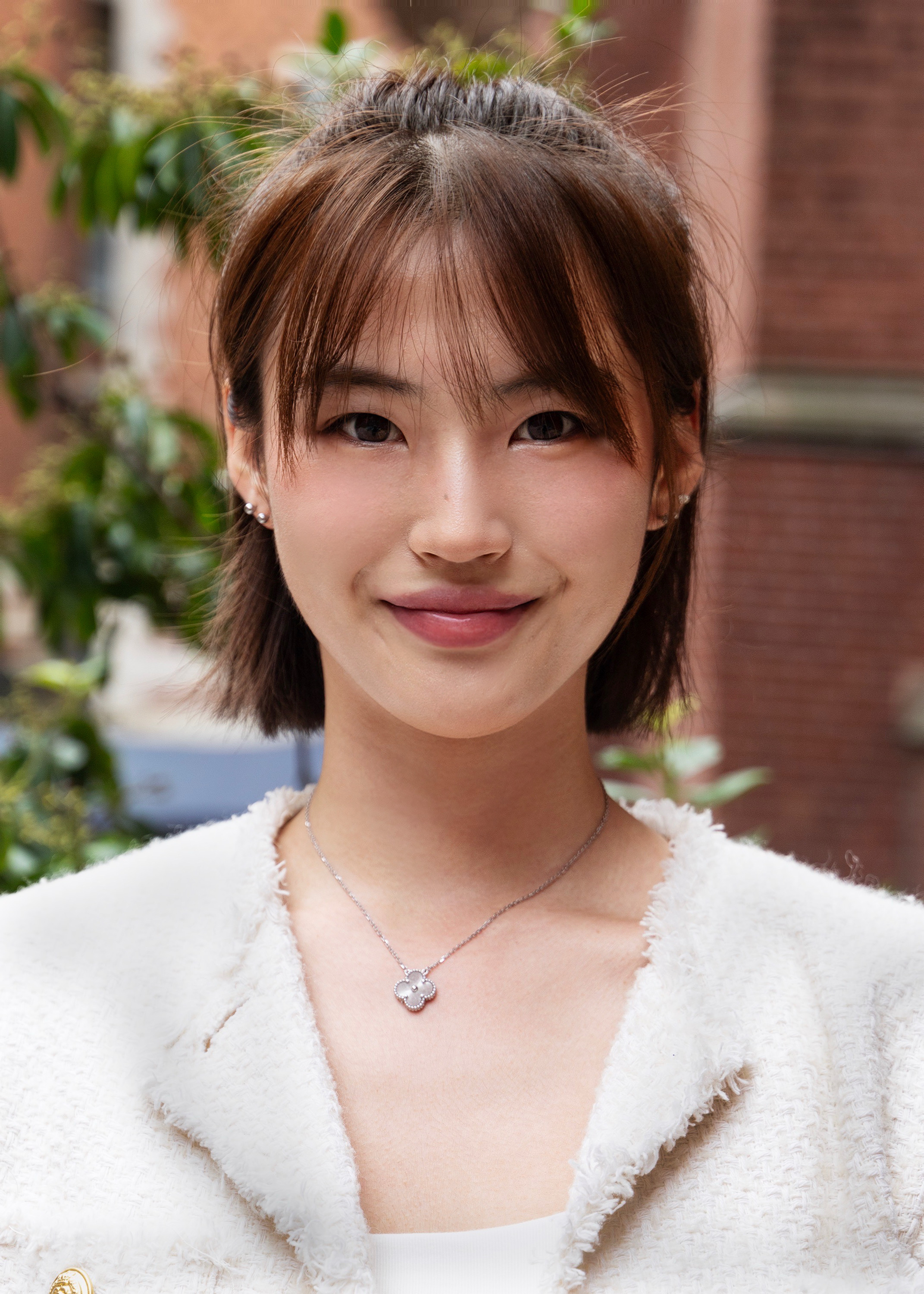Zoe is posing for a photo. She has short, straight, warm brown hair coming down to her chin. She is smiling and looking at the camera. She is wearing a fuzzy, off-white blazer jacket.