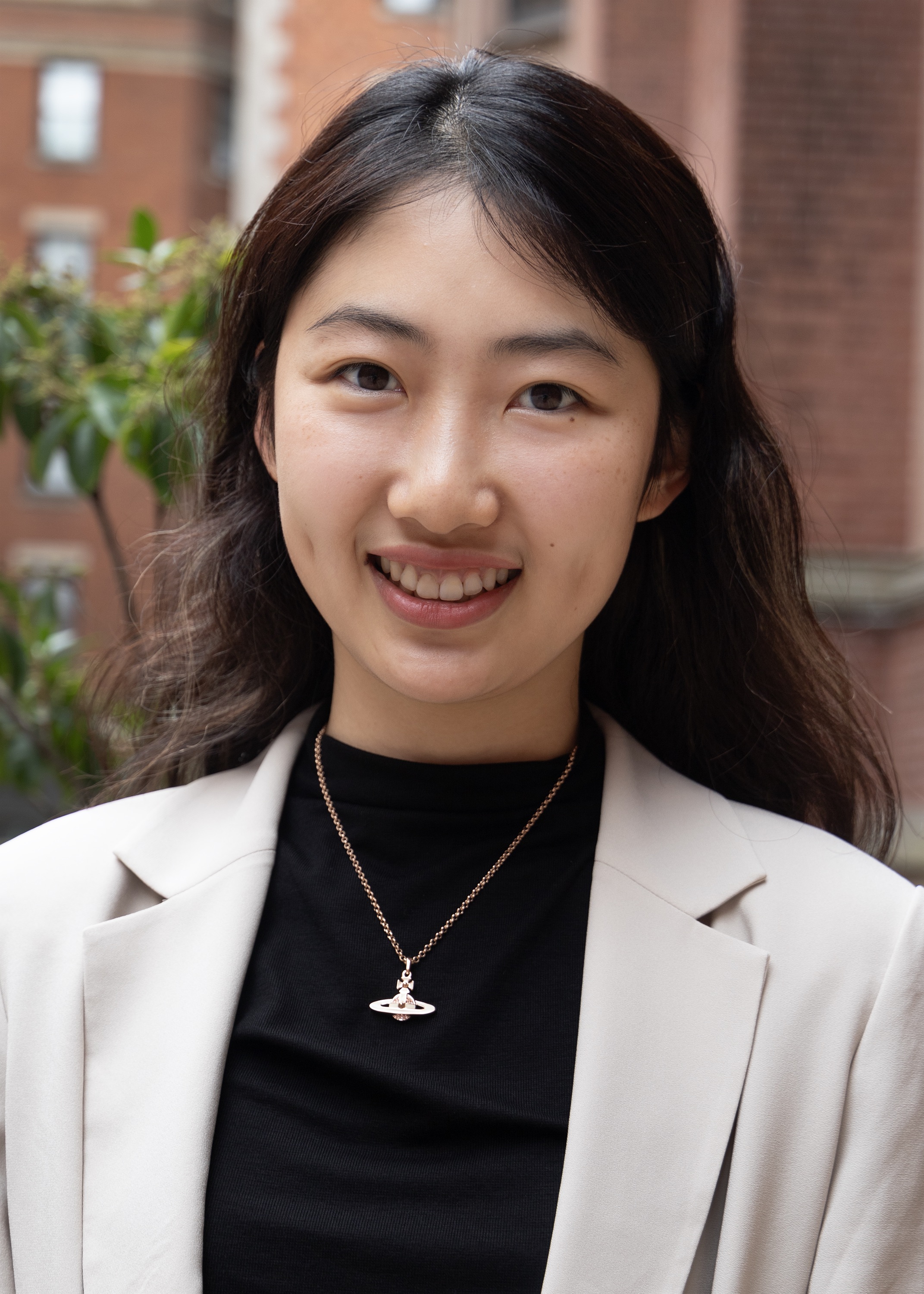 Xinyi is smiling for a photo. She has long, dark brown hair in loose waves falling behind her shoulders. She is wearing a black shirt and an off-white blazer.