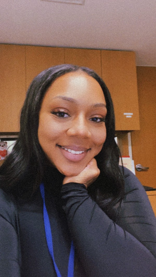 Smiling woman with long dark hair wearing a black log sleeve top and blue lanyard, seated in an office